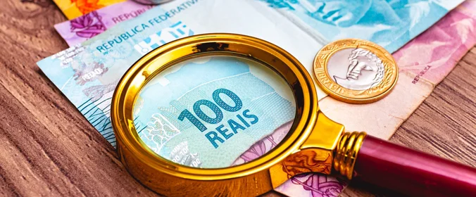 foto de cédulas do real brasileiro, com uma moeda em cima e uma lupa maximizando o valor da nota